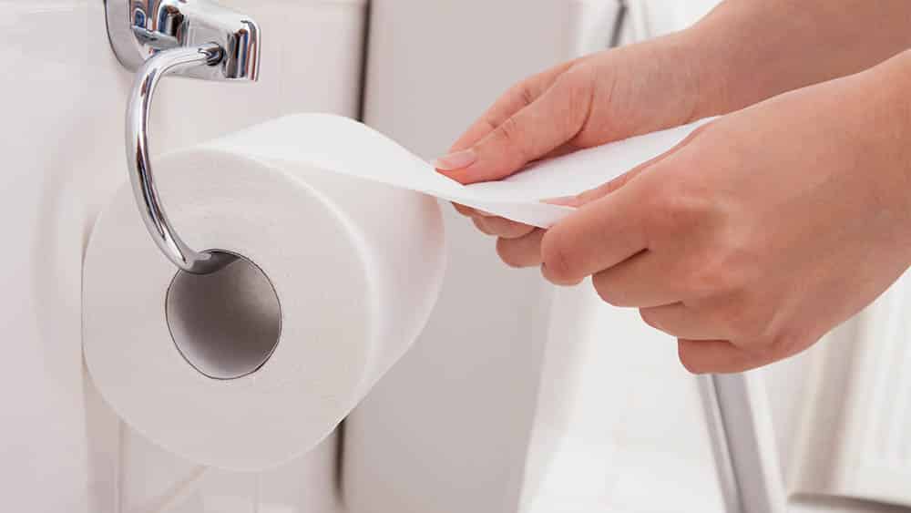 Close nas mãos de uma mulher desenrolando papel higiênico em um banheiro.