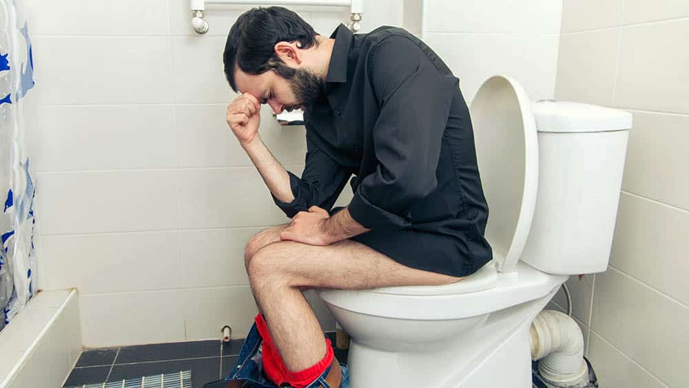 Homem de perfil sentado no vaso sanitário com a mão na cabeça e expressão de dor.