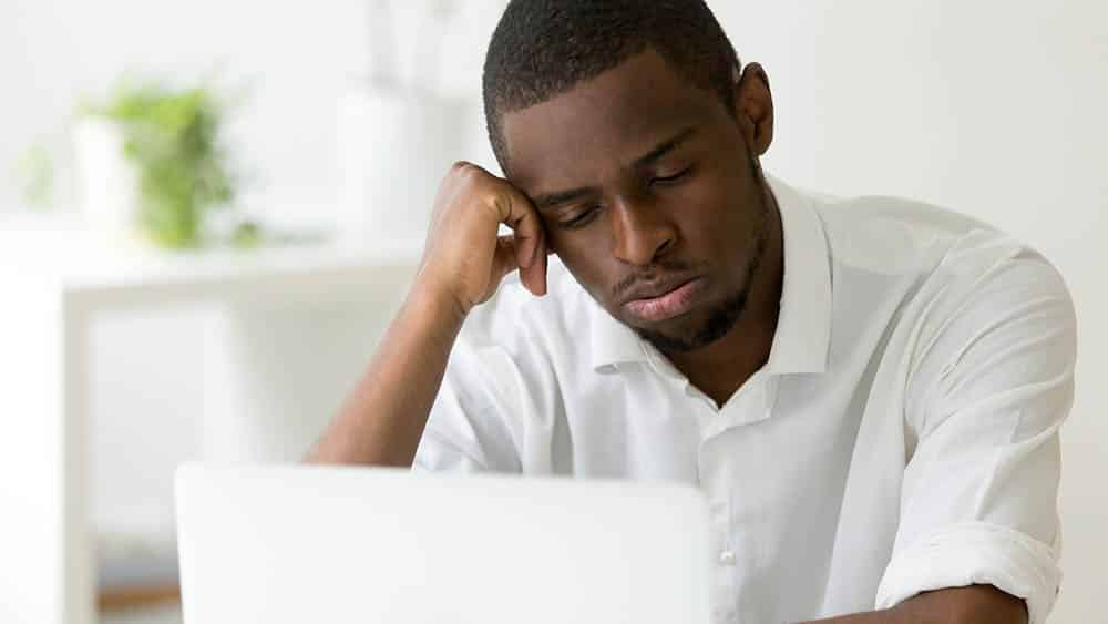 Homem em frente a um notebook com a cabeça apoiada em uma das mãos expressando tédio.