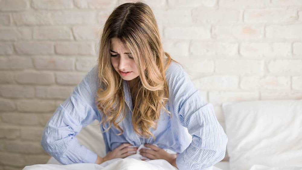 Mulher sentada na cama pressionando o abdômen com as mãos e expressão de dor.