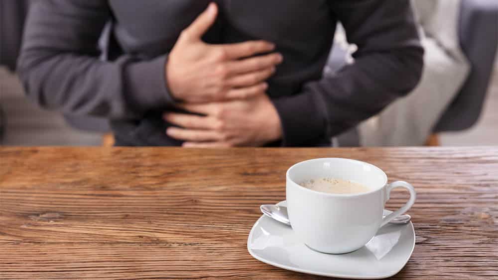 Homem pressionando estômago ao fundo e xícara de café em primeiro plano.
