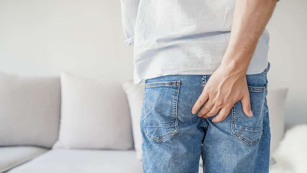Homem de costas de calça jeans com a mão nas nádegas indicando coceira no ânus.