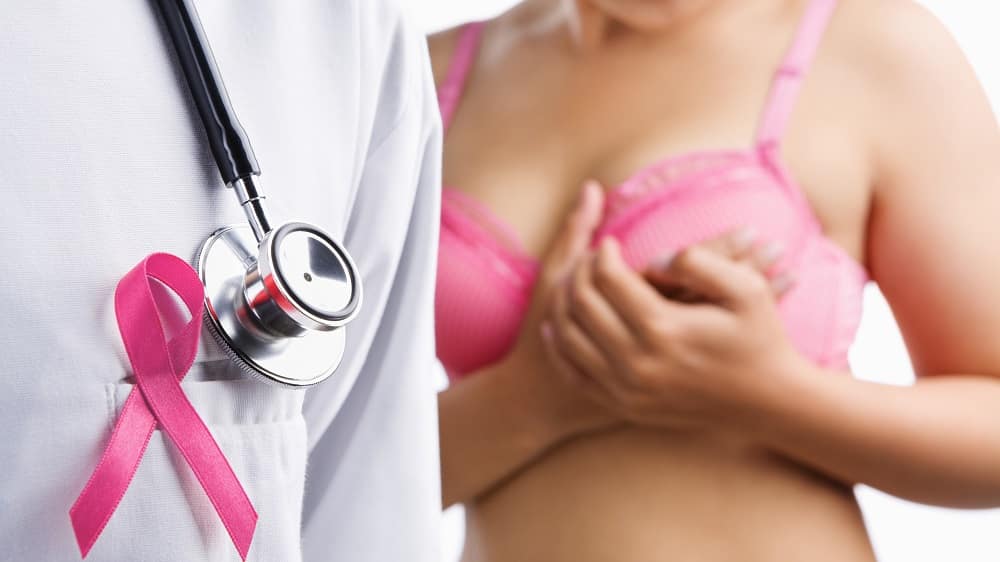Médico com o laço rosa que representa a luta contra o câncer de mama, enquanto mulher toca o próprio seio