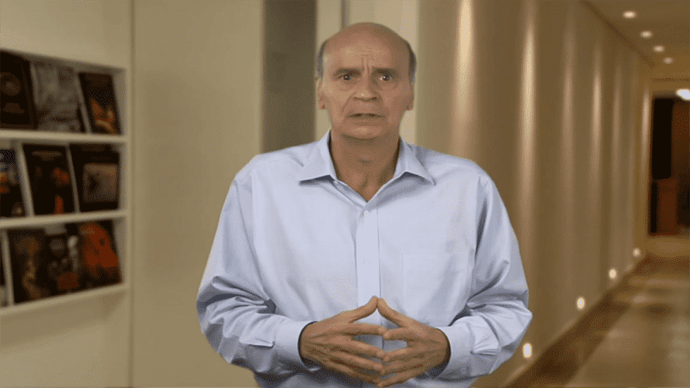 Drauzio Varella de mãos cruzadas em um corredor falando sobre tumores
