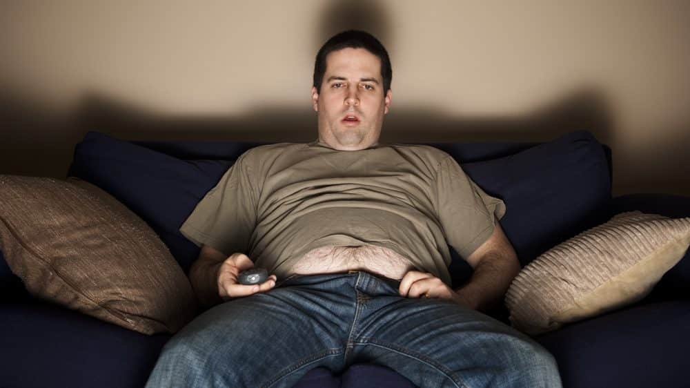 homem sedentário e com sobrepeso sentado no sofá, assistindo à TV