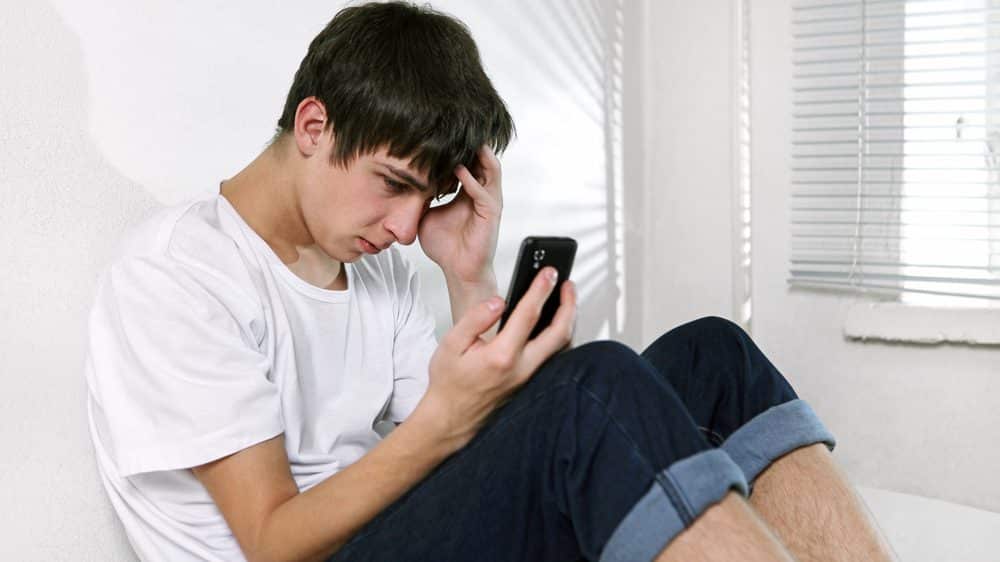 adolescente triste, sentado na cama, com celular na mão. solidão crônica interfere na saúde