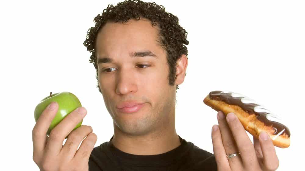 rapaz segura maçã em uma mão e bomba de chocolate em outra. há relação entre obesidade e dieta