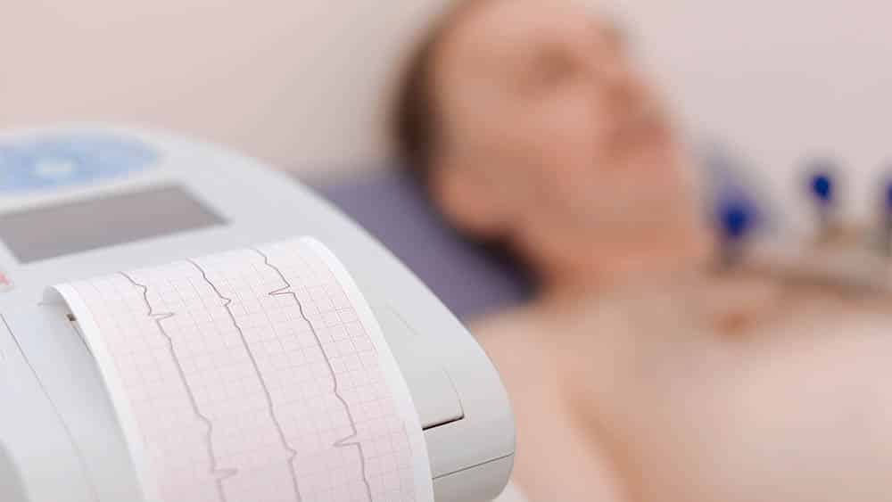 Aparelho emitindo eletrocardiograma com paciente deitado desfocado ao fundo.