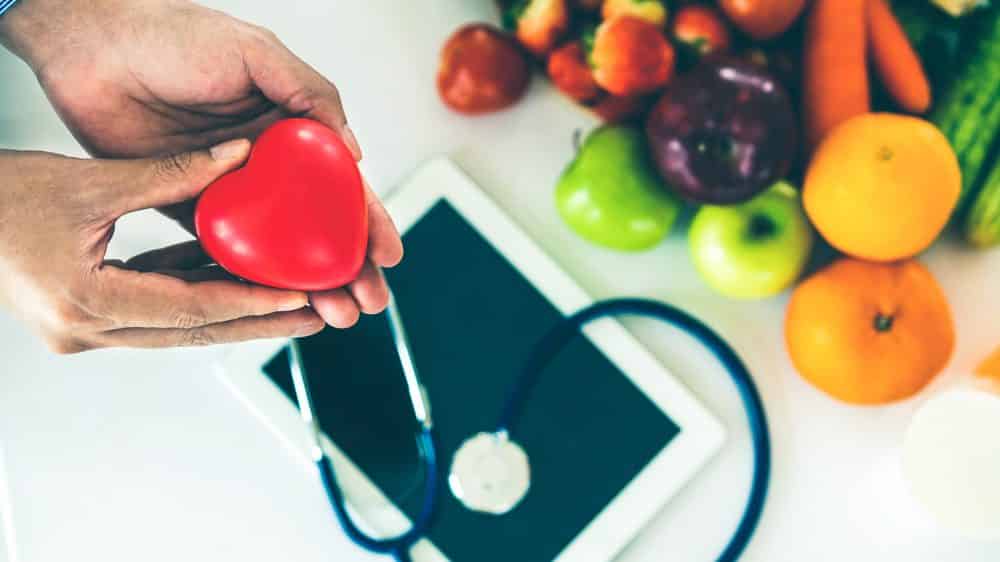 homem segura modelo de coração com frutas, esteto e ipad ao fundo. Há controvérsias sobre o colesterol entre médicos