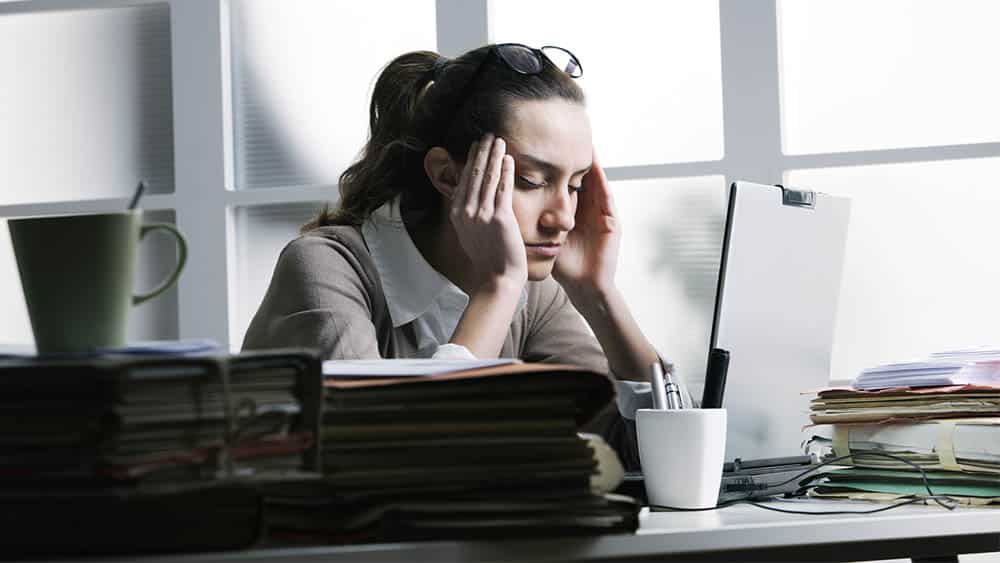 Executiva em frente a um notebook com as mãos nas laterais da cabeça e olhos fechados indicando dor.
