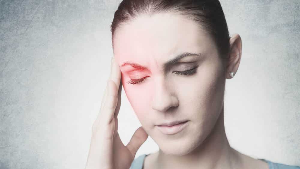Mulher com mão na lateral da cabeça e olhos fechados indicando enxaqueca.