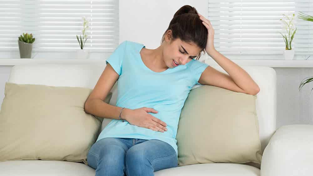 Mulher sentada em um sofá com a mão no abdômen e expressão de dor.