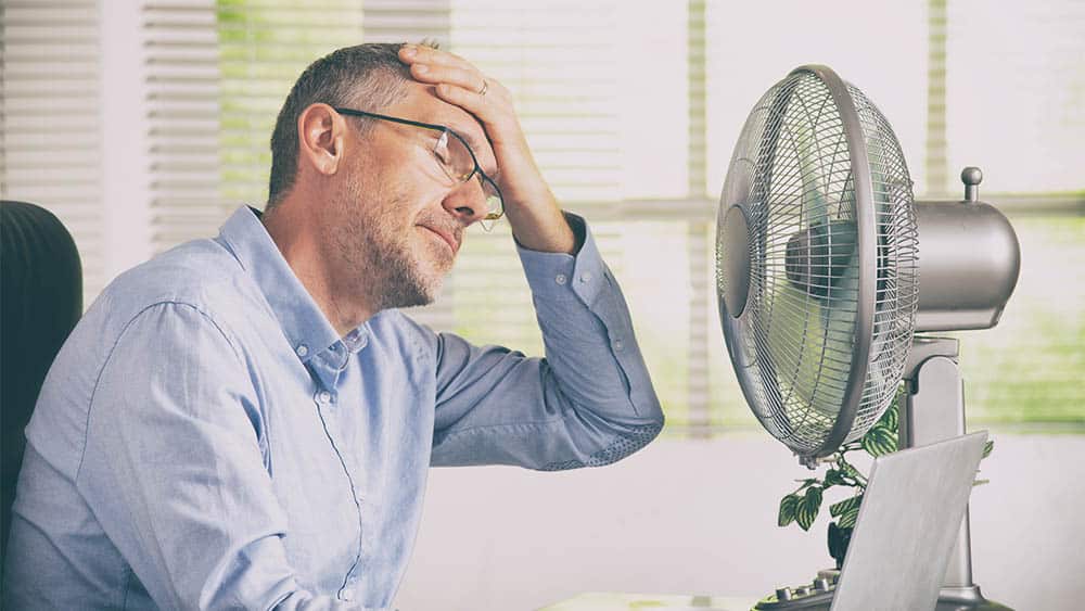Homem em escritório com mão na testa em frente a ventilador.