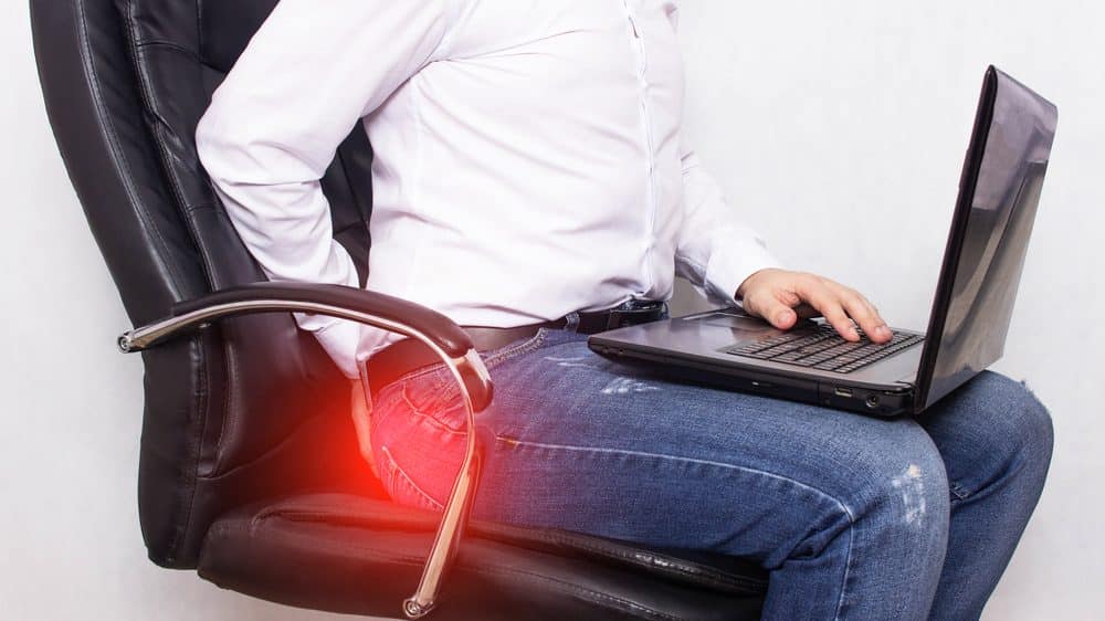 homem sentado, segurando computador no colo, com sinal de dor no ânus. Ficar muito tempo sentado aumenta risco de hemorroidas.