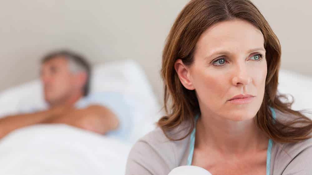 Mulher triste com marido deitado na cama desfocado ao fundo.