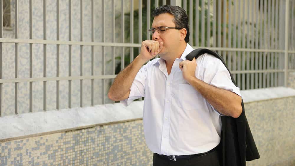 Homem tossindo com mão na boca enquanto caminha pela rua.