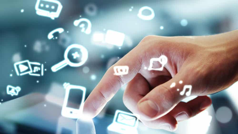 mão toca tablet e ícones de redes sociais saltam da tela. Internet divulga desinformação em excesso