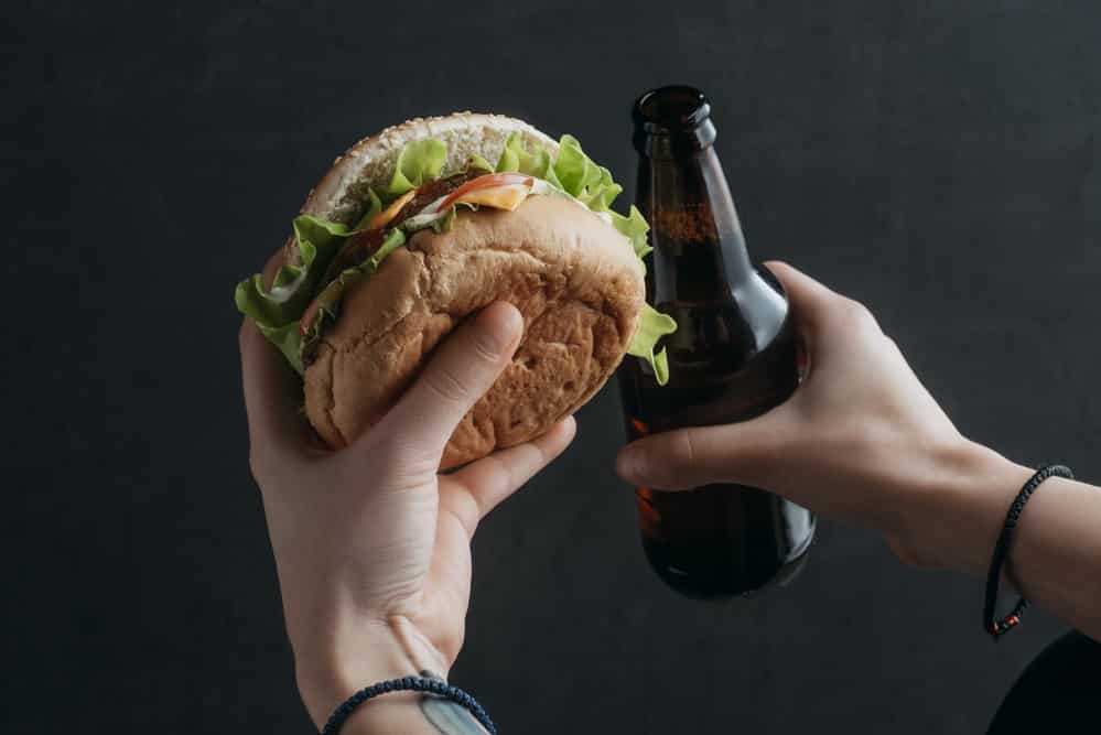 homem segura hambúrguer e cerveja, mostrando estilo de vida prejudicial à saúde