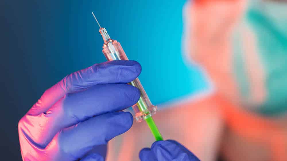 Profissional de saúde com máscara e luva segurando uma seringa.