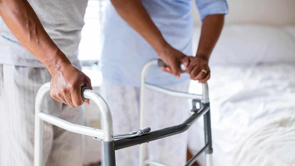 Homem apoiado em um andador sendo ajudado por um profissional de reabilitação.