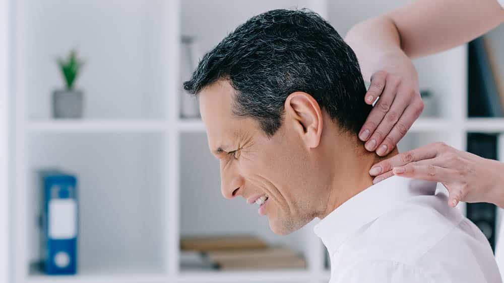 Homem de perfil com expressão de dor de torcicolo enquanto a região cervical é massageada.