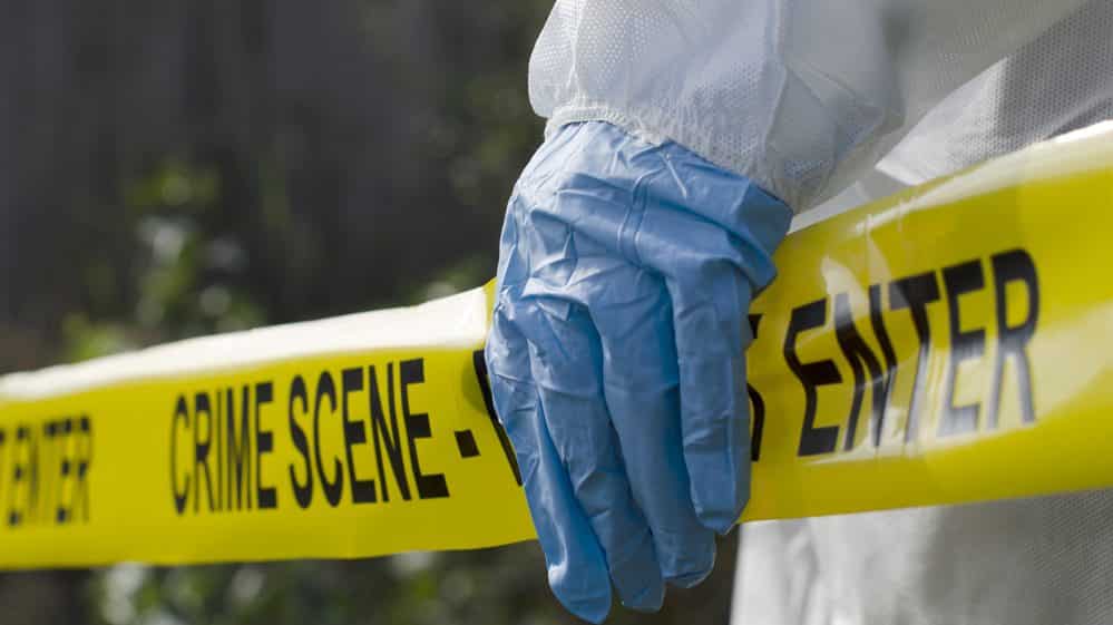 Mão com luva sobre uma faixa amarela típica de isolamento de cenas de crime.