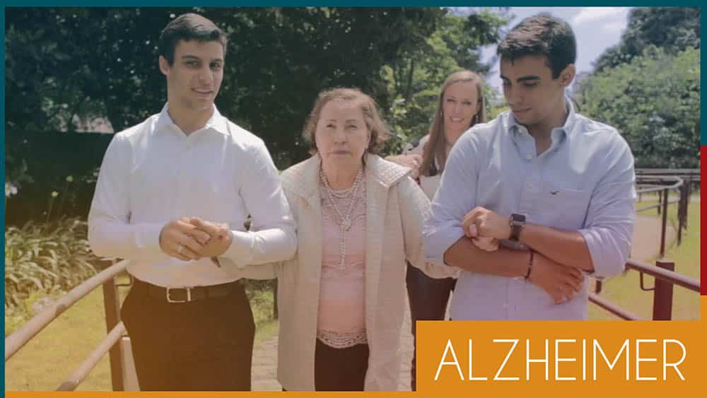 Dona Marisa, paciente com Alzheimer, caminha auxiliada por cuidadores.
