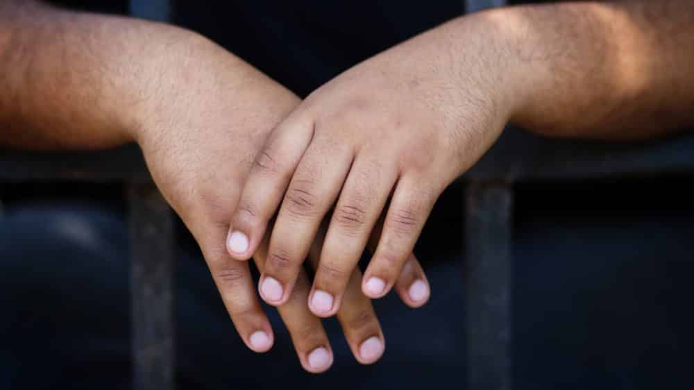 Mãos de rapaz preso atrás de grades.