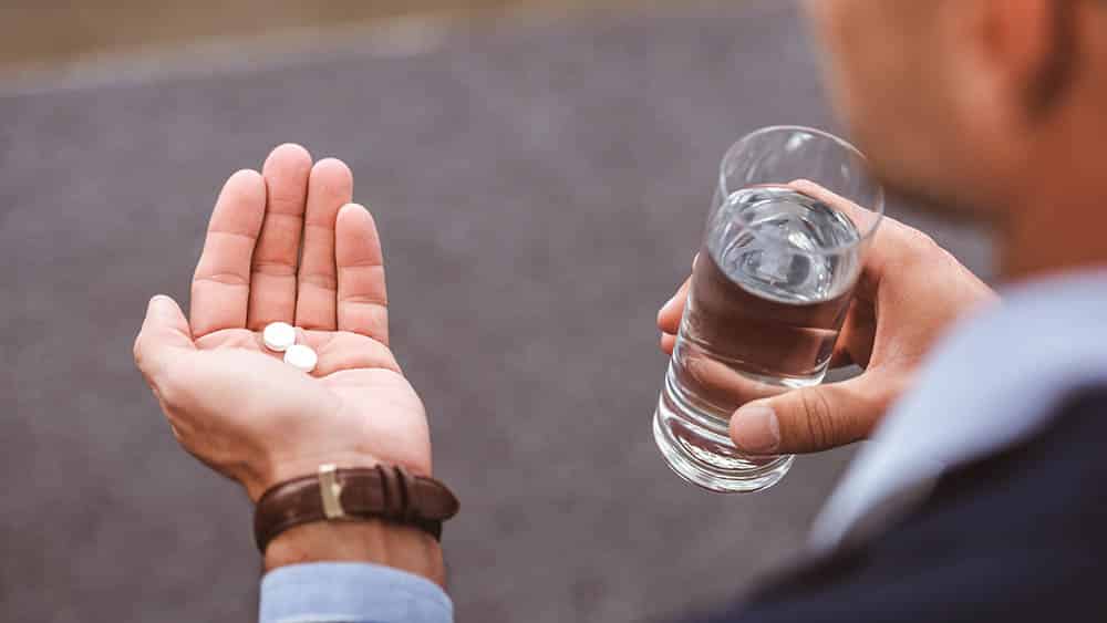 Executivo com um copo de água em uma mão e dois comprimidos brancos em outra prestes a ingeri-los.