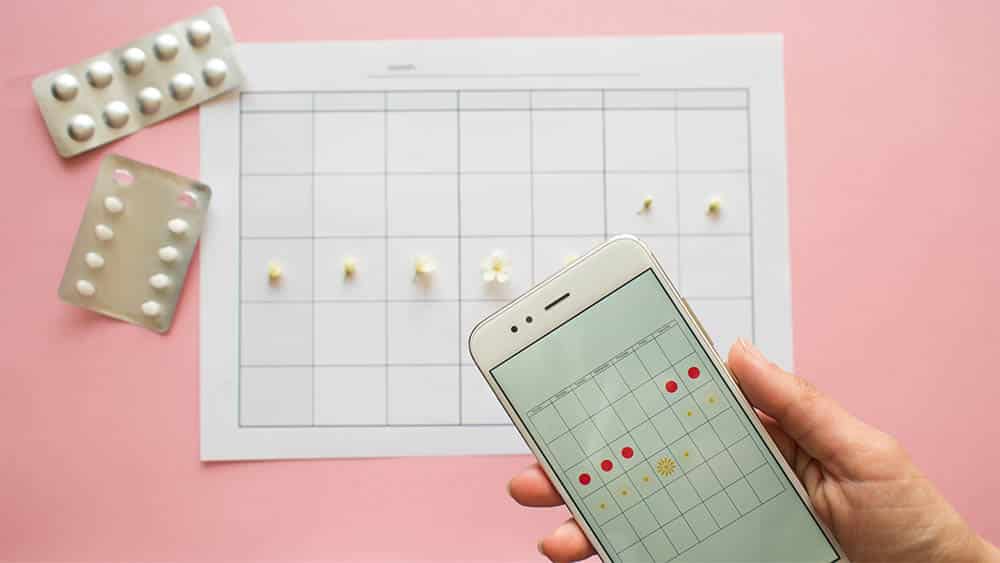 Vista de cima de calendário com dias marcados, cartelas de anticoncepcional e, em primeiro plano, mão feminina segurando celular com aplicativo de ciclo menstrual na tela.