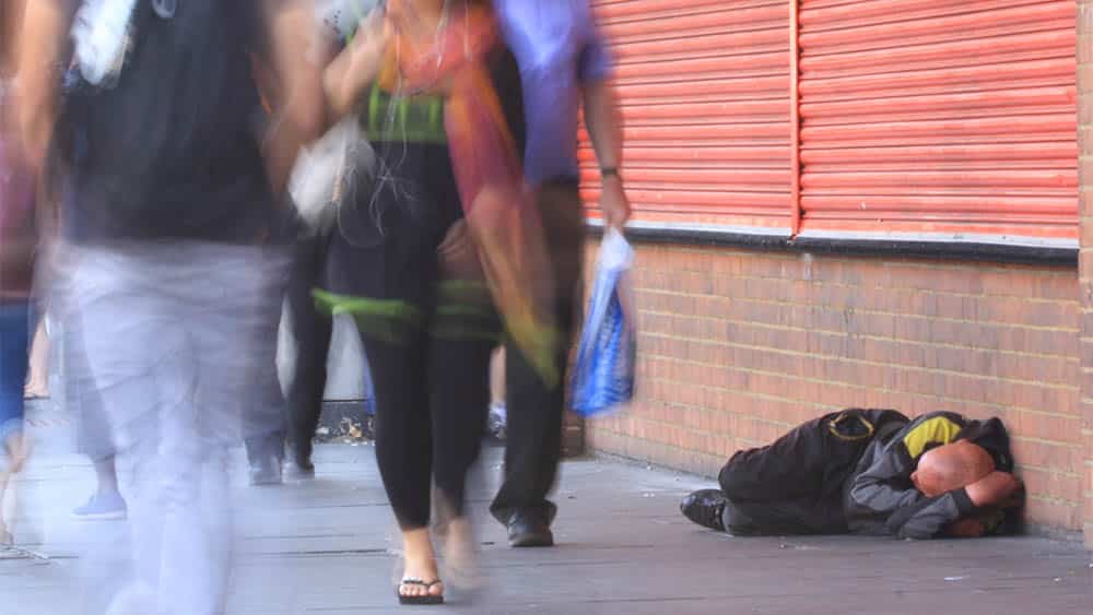 Atendimento a pessoas em situação de rua inclui necessidades básicas Homem em situação de rua deitado em calçada com pessoas passando ao largo.