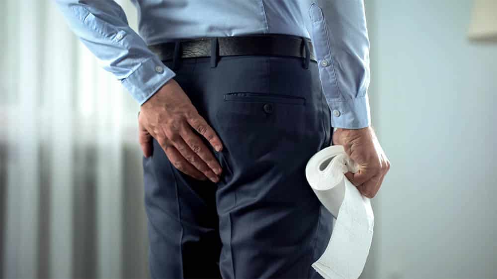 Executivo de costas com mão nas nádegas e segurando rolo de papel higiênico, em sinal de fissura anal
