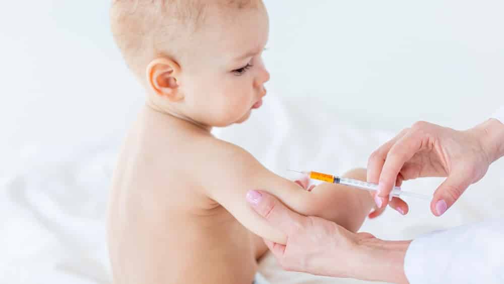 Bebê sentado e mãos de mulher segurando seu braço antes de aplicar vacina. Bebês tomam grande quantidade de vacinas.