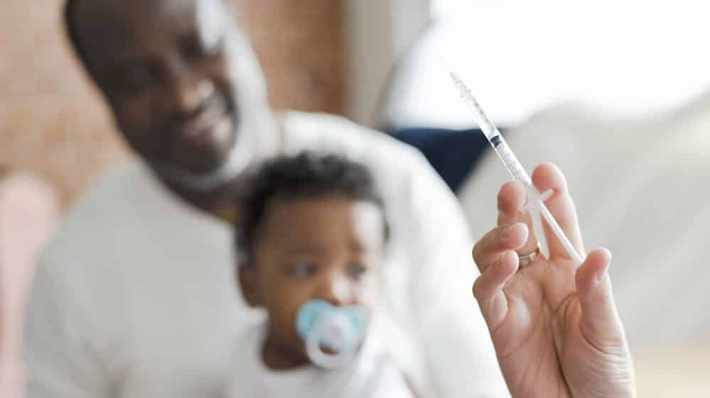 Começa a Campanha Nacional de Vacinação contra o Sarampo bebê no colo do pai ao fundo. em primeiro plano, mão segurando injeção.começa campanha de vacinação contra o sarampo