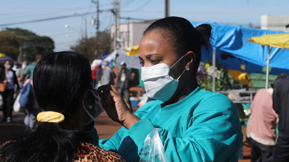 mulher de máscara ajusta a máscara de outra mulher em feira livre no Paraná. A epidemia brasileira atinge todos os estados
