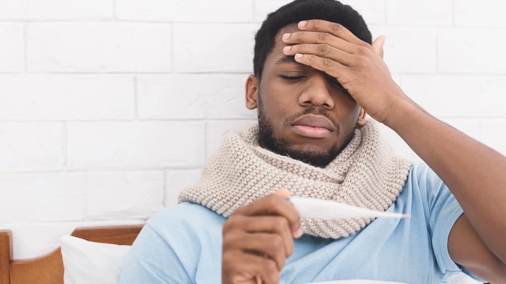 homem doente na cama olha termômetro. febre é um dos sintomas da covid-19