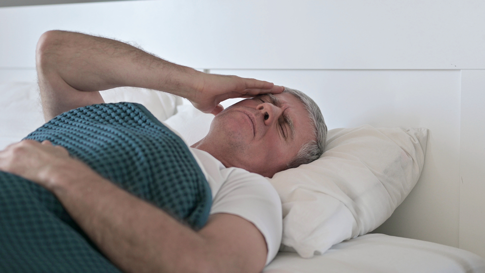 homem idoso de cama, com mão na cabeça, em sinal de dor. Sequelas da covid são variáveis