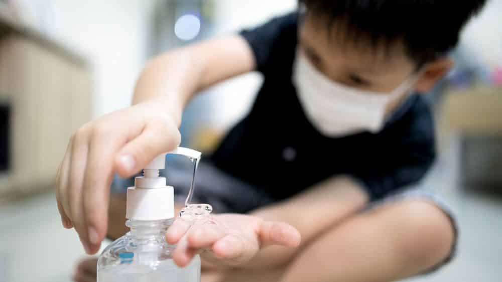 menino pequeno de máscara passa álcool gel nas mãos. Intoxicação por álcool gel tem aumentado
