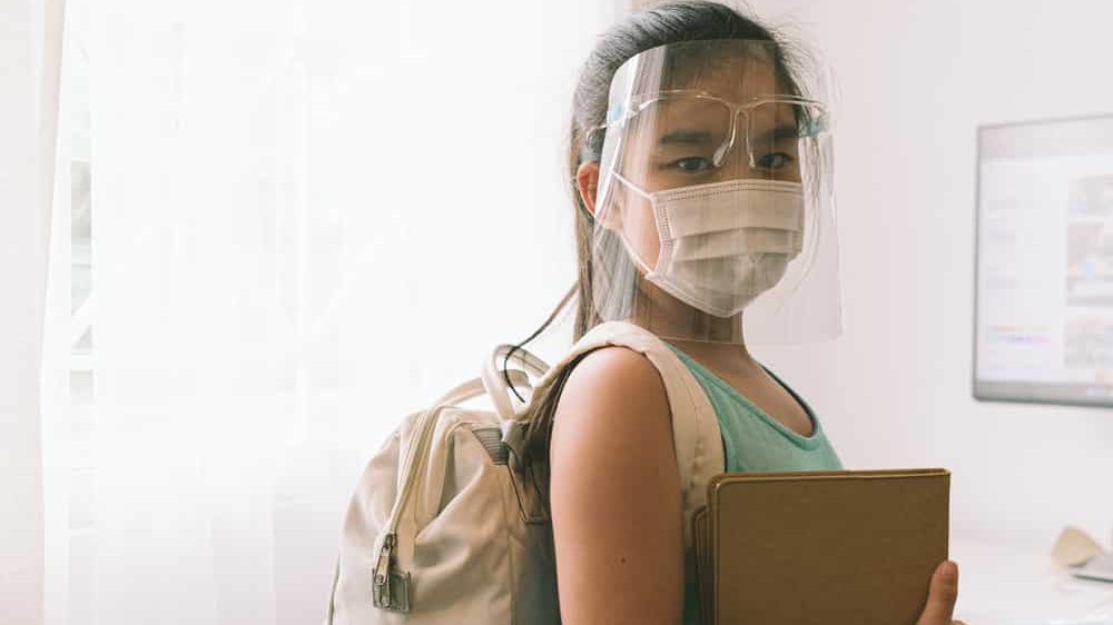 menina de máscara e proteção facial carrega mochila e caderno para a volta às aulas na pandemia