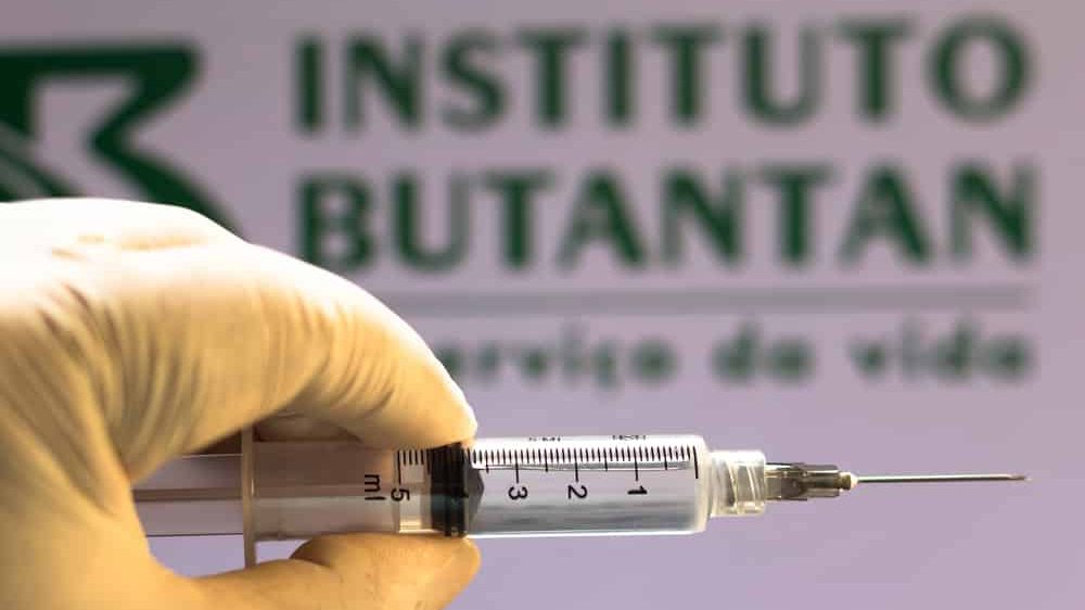 mão enluvada segurando seringa. AO fundo, lê-se Instituto Butantan, que produzirá CoronaVac